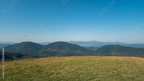 Obraz na płótnie High-angle panoramic view of rolling mountain ridges, sloping grassy hillside, and expansive clear blue sky on a sunny day