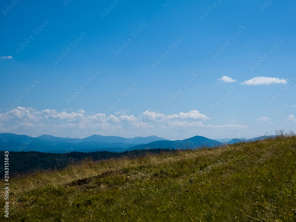 Obraz premium Wide outdoor stock photo of a narrow dirt trail stretching across a grassy alpine mountain ridge toward distant blue peaks under a vast clear blue sky. Concept for trekking journey, eco adventure, tra