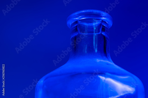 Close-up of a clear glass bottle neck and upper shoulder centered against a solid blue background. Highlights and reflections are visible on the glass surface.