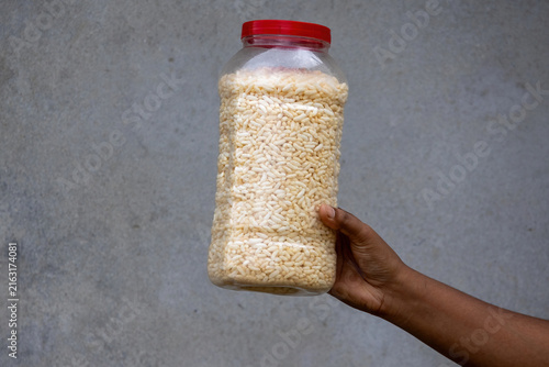 Person holding plastic container filled with crispy puffed rice against rustic grey wall background. Traditional South Asian muri stored for daily tea snack.