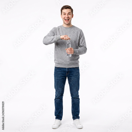 Full-length portrait of a happy young man in casual clothes opening a plastic water bottle, standing on a white background.