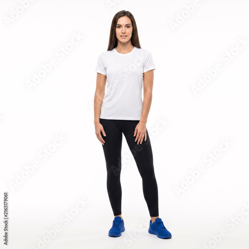 Full-length studio shot of a fit young woman in a white t-shirt, black leggings, and blue athletic shoes, standing against a white background.