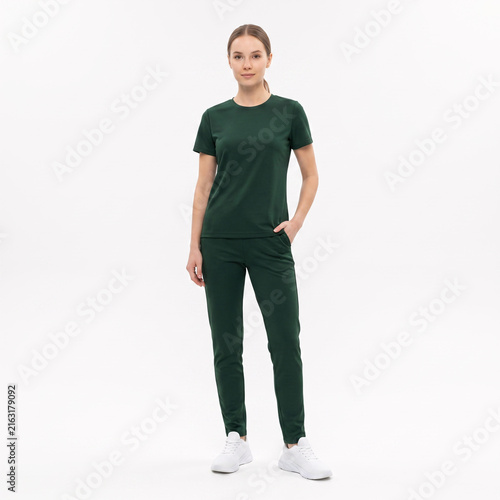A young woman stands confidently in a full-body shot, wearing a dark green short-sleeved t-shirt and matching slim-fit pants, paired with white sneakers, against a clean white background.