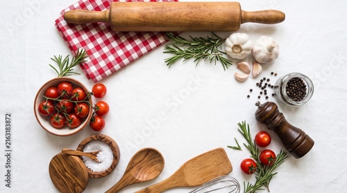 “Flat lay of cooking ingredients and utensils on white marble surface, circular arrangement with tomatoes, rosemary, garlic, salt, pepper, rolling pin, spoons, and cloth.”