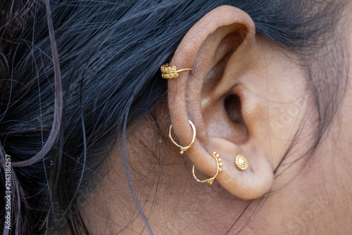 Female ear adorned with multiple traditional golden rings and stud piercings. Ornate gold jewelry worn for traditional beauty and fashion accessory.