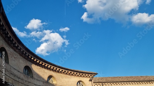 Historic curved brick architecture with arched windows beneath a vivid blue sky and white clouds, viewed from a courtyard.