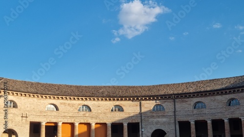 Historic curved brick architecture with arched windows beneath a vivid blue sky and white clouds, viewed from a courtyard.