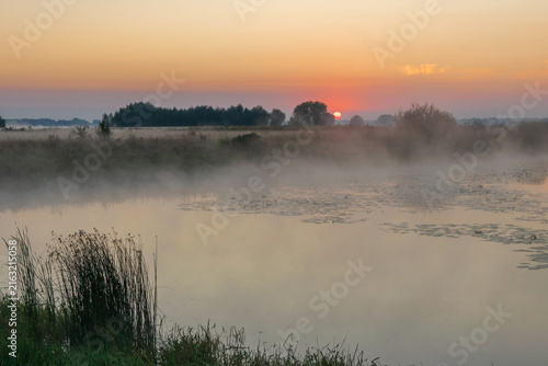 Принти на полотні A misty river flows gently under a warm orange sunrise, with hazy trees and gras