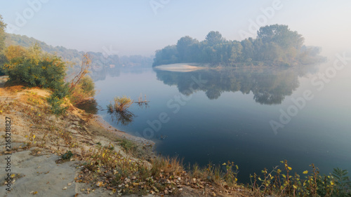 A serene, misty morning landscape features a calm river reflecting lush green trees and a small island under a hazy sky