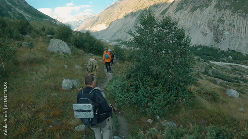 Group hiking mountain path surrounded by greenery and rocky slopes. Sunlight highlights trail and distant peaks. Mood is adventurous and serene. Ideal for travel, outdoor activity, or nature themes