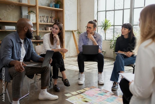 Diverse group of young professionals sitting in a circle during a collaborative workshop or brainstorming session in a bright, modern office space with sticky notes on the floor