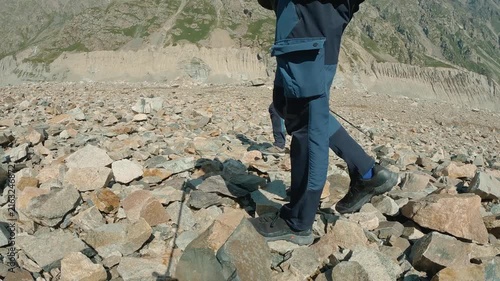 Low angle view of hikers in outdoor gear stepping over rocky mountain path. Bright daylight highlights textures of stones distant alpine slopes. Evokes adventure, endurance, nature exploration