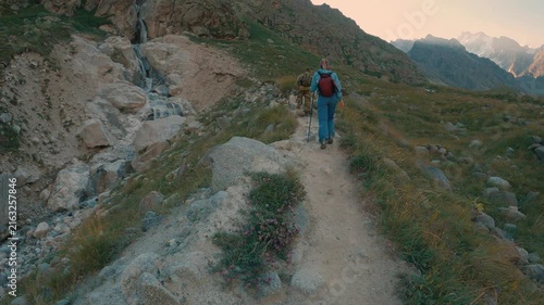 Hiker in blue outfit red backpack walks dirt trail beside rocky stream small waterfall. Distant snowcapped mountains under soft golden light. Wildflowers grasses line path