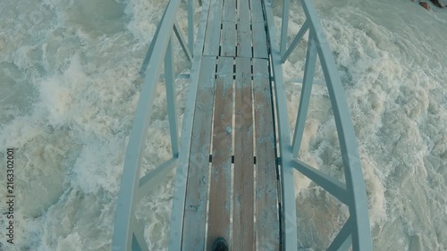 First person view walking across narrow wooden bridge spanning turbulent river. Metal railings frame path. Rough water flows fast around large rocks. Cool tones dominate