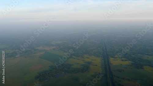 Drone flight showing green fields, agricultural farmland and forest line vanishing into hazy horizon during soft morning light.