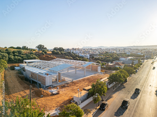 Aerial view of a construction site featuring a steel frame structure on a dirt lot, adjacent to a road with moving cars, with the urban landscape of Chania, Greece.