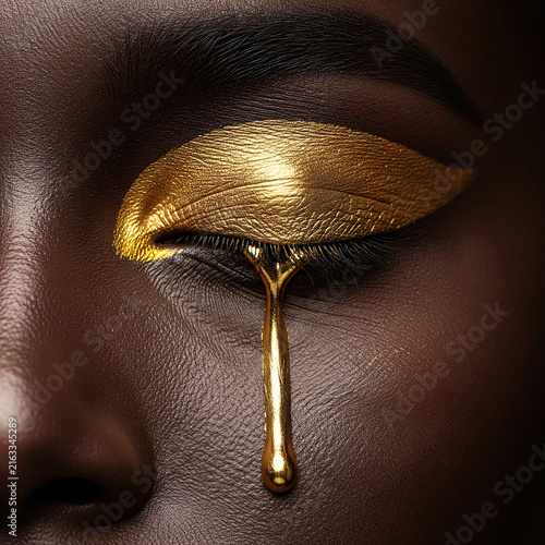 Close up macro beauty portrait of dark skin woman with metallic gold eyeshadow and liquid gold tear dripping down cheek. High fashion avant garde creative makeup, artistic luxury cosmetic concept.