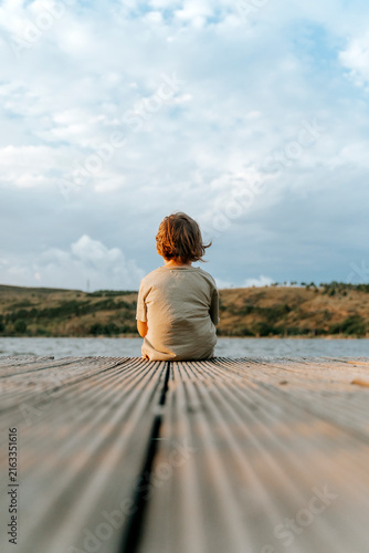 фотографія Lonely child sitting on a wooden dock looking out at a calm lake