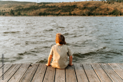 Lonely child sitting on a wooden dock looking out at a calm lake