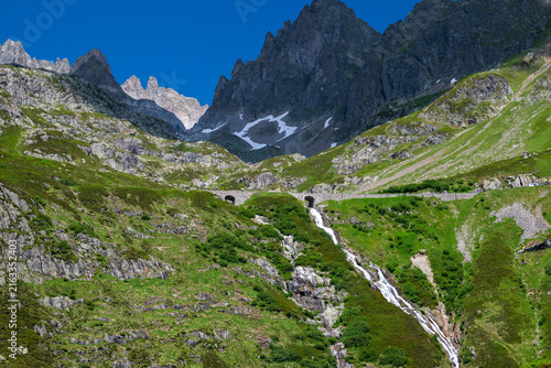 Quadro su tela Mountain stream cascading through green alpine slopes beneath rugged snow-dusted peaks in the Swiss Alps