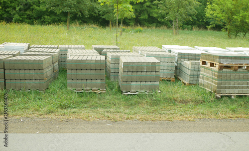 Concrete paving blocks stacked interlocking pavers rectangular, bricks curb blocks stacked wooden pallets green plastic banding straps strapping, stored outdoors grassy verge and a paved road stored