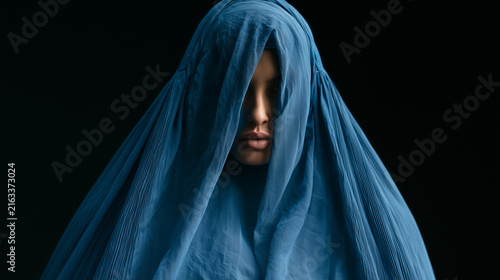 Woman wearing a traditional blue burqa standing against a dark background, symbolizing culture, identity, and the complex layers of tradition and privacy. Afghan culture, burqa por
