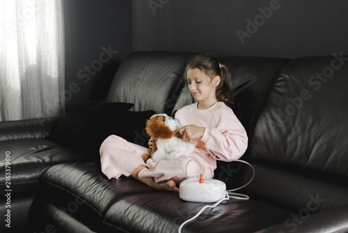 A little girl sitting on a black leather sofa at home, holding a stuffed toy and using a nebulizer inhalation mask for respiratory therapy. Child smiling while receiving asthma or cold treatment