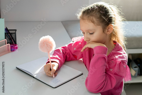 A focused little girl sits at the desk and carefully writing in her notebook while studying at home during an online class on a tablet. Concept of school, homework and the beginning of the school year