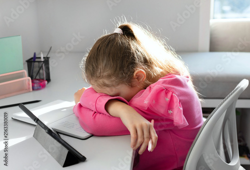 A young girl sits at a desk, head down, appearing deeply exhausted and unable to proceed with her studies during an online lesson. Concept of school, homework and the beginning of the school year