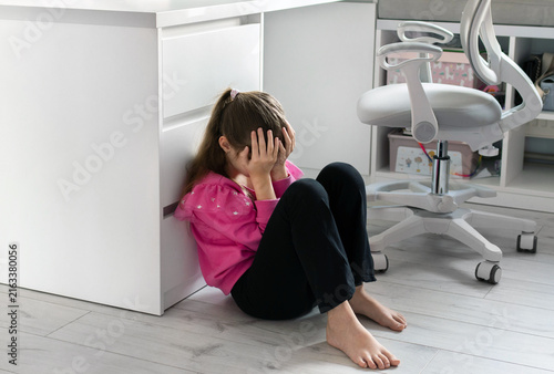 A little girl sits on the floor clutching her head, reflecting distress from a misunderstanding with her parents, a family conflict, problems at school or misunderstanding with friends