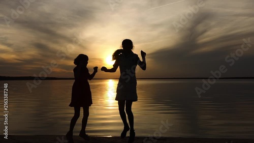 Two little sisters jumping happily in front of the setting sun and the big lake. Beautiful sunset.