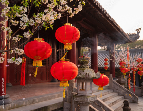 Red Chinese lanterns hanging from tree branches with white flowers, as traditional Asian decorations adorn an outdoor temple, creating a festive atmosphere for celebration and culture.
