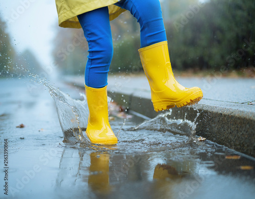 A child's feet in yellow rain boots splashing in a water puddle.