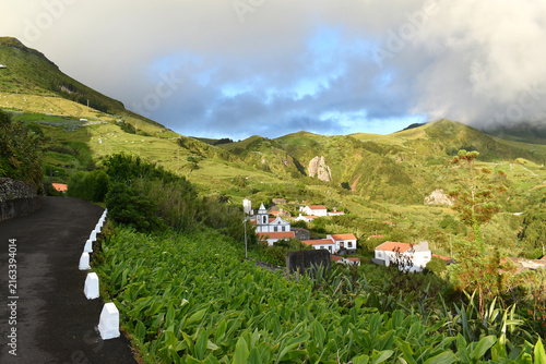 The picturesque village of Lajedo on the island of Flores, Azores, nestled among green hills amidst low-hanging clouds and a sunset, bordered by a road with white marker posts
