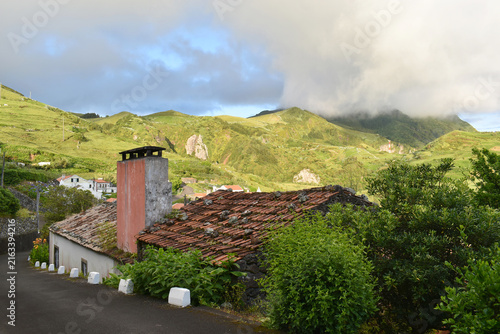 The picturesque village of Lajedo on Flores Island, Azores, nestled among green hills beneath low-hanging clouds at sunset; in the foreground, a roof with clay tiles weighed down by stones