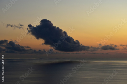 A dark cloud over a calm ocean at sunset, with the reflection of the shadow on the water's surface
