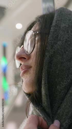 Young woman with long dark hair and round glasses wears. Hand maintains grip on fabric suggesting minimal. Background features soft lighting and occasional colored lights