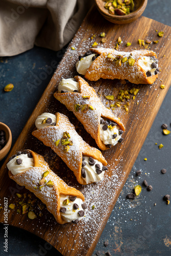 Traditional Sicilian cannoli pastries filled with ricotta cream, chocolate chips and crushed pistachios on a wooden board. Delicious sweet Italian dessert close up