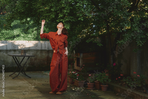 Photography Smiling woman in a red jumpsuit stands in a sunlit garden, reaching upward with one arm