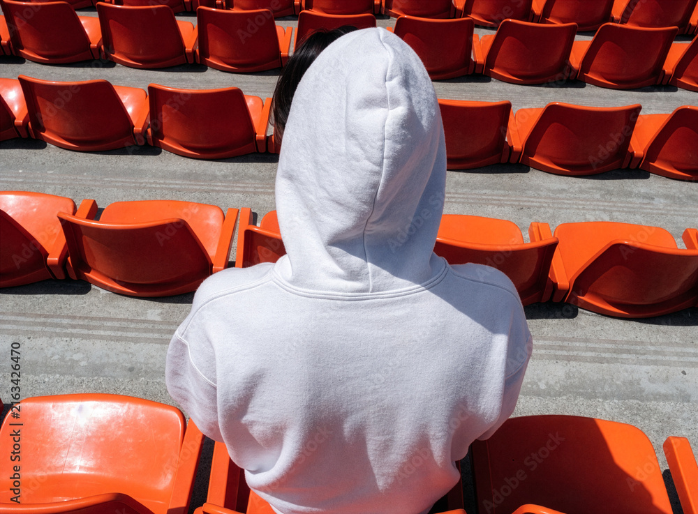 Fototapeta premium Person in white hoodie sitting in empty orange stadium seats outdoors