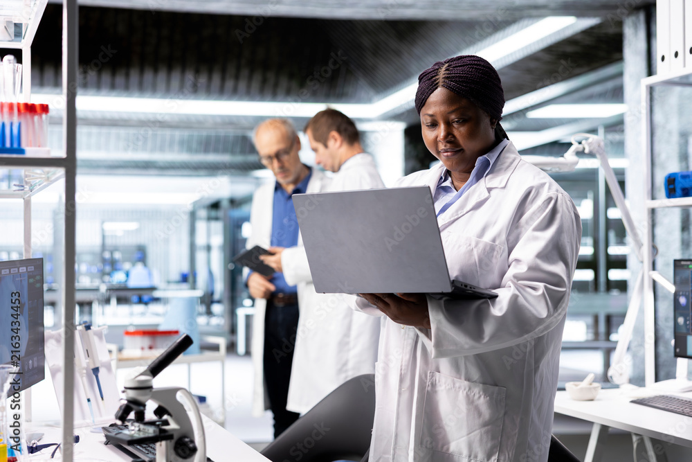 Fototapeta premium Black female researcher in a clinical research lab studies experiment data with a laptop, showcasing biotechnology, genetics research and pharmacology for progress in healthcare and medical research.