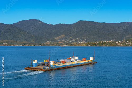 Car ferry vessel navigates through deep blue sea waters with vehicles on deck, set against a backdrop of lush green coastal mountains and a clear, bright blue sky