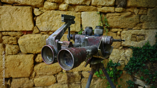 Vintage military observation binoculars mounted on tripod against old rustic stone wall