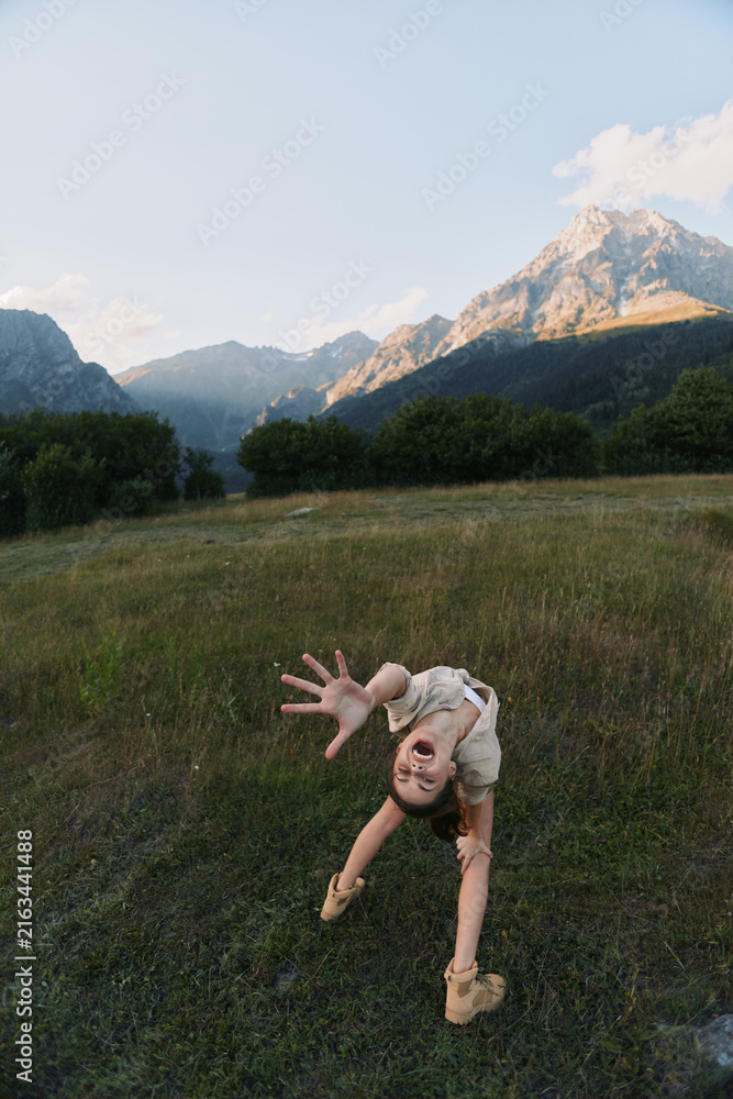 Naklejka premium Energetic woman bends toward the camera in a sunlit meadow, mountains in the background, casual outdoor wear, capturing a playful and adventurous lifestyle moment
