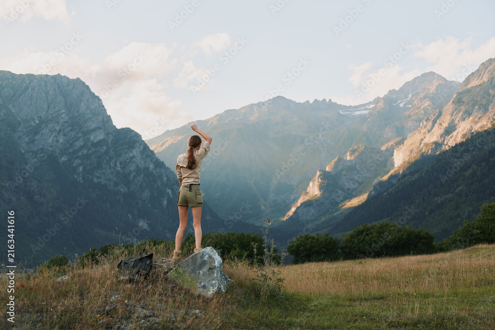 Naklejka premium Active woman stands on a rock in a mountain landscape, embracing outdoors life amid rugged peaks and blue sky, evoking hiking, nature, and scenic exploration.