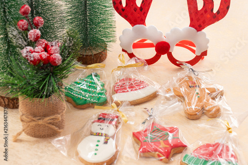 Individually wrapped christmas homemade cookies in various festive shapes. Placed on a light surface with small decorative christmas trees and playful reindeer antler glasses