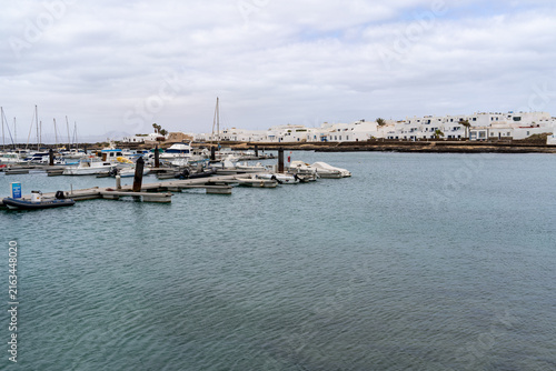 The serene harbor of La Graciosa, Lanzarote, is filled with boats, docks, and white buildings under a cloudy sky, creating a peaceful scene.