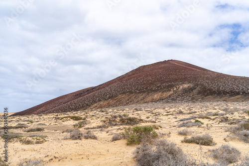 A scenic view of a volcanic mountain on La Graciosa island, under a cloudy sky with arid landscape.