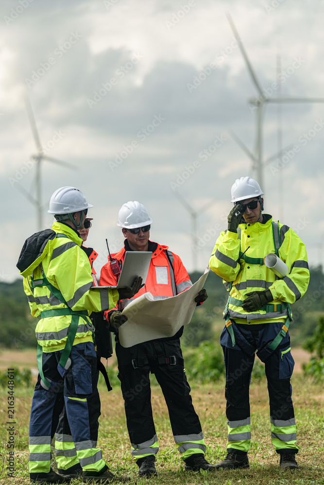Fototapeta premium A mixed team of wind farm engineers in yellow and orange hi vis walk the meadow while consulting the site plan on a green meadow with rows of turbines behind in bright overcast daylight.