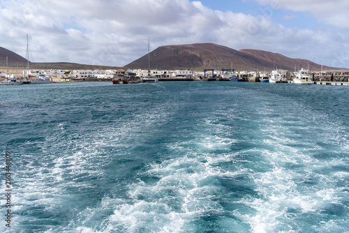 A scenic view of La Graciosa, with the wake of a boat leading towards the harbor. The island's mountains are visible in the background.